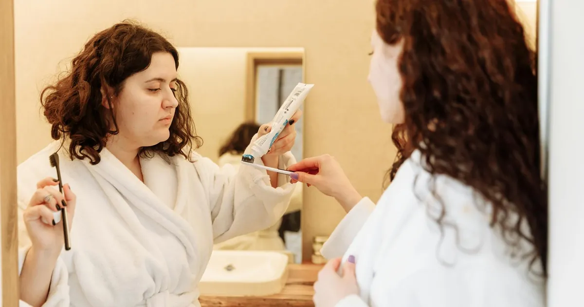 Two women in bathrobes share a morning routine, applying toothpaste at a bathroom sink.