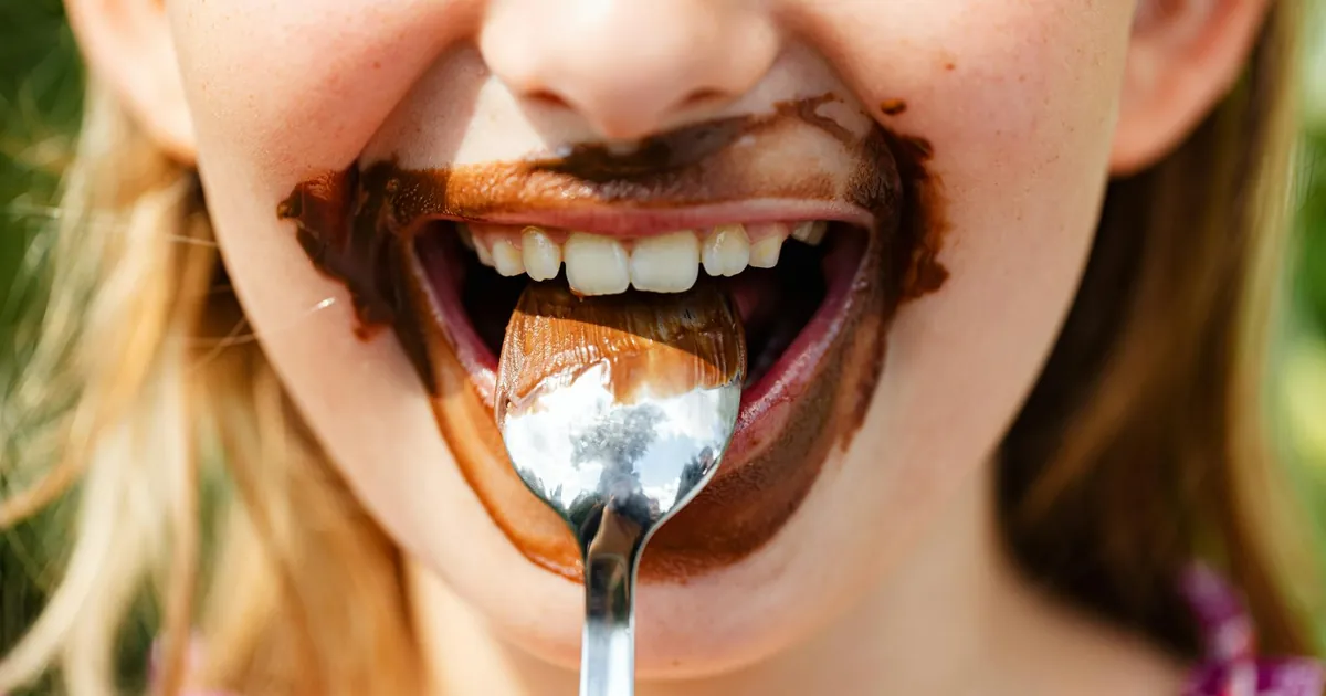 Close-up of a child's face enjoying a messy chocolate treat, smiling with a spoon.