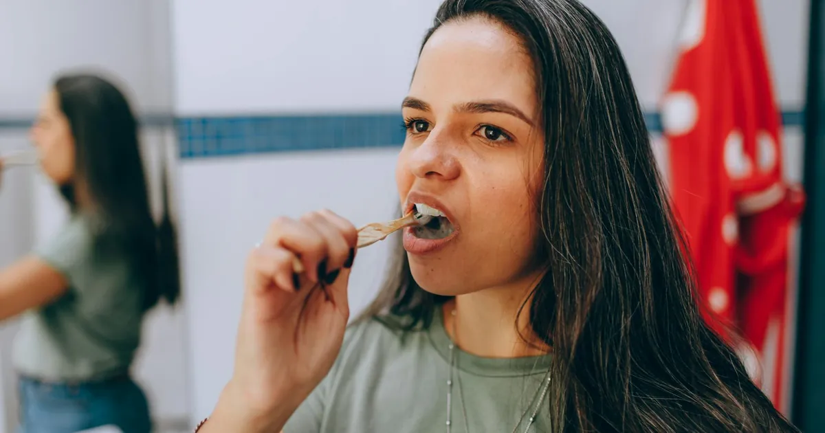 Woman brushing her teeth using a natural toothbrush in a bathroom, promoting oral hygiene and sustainability.