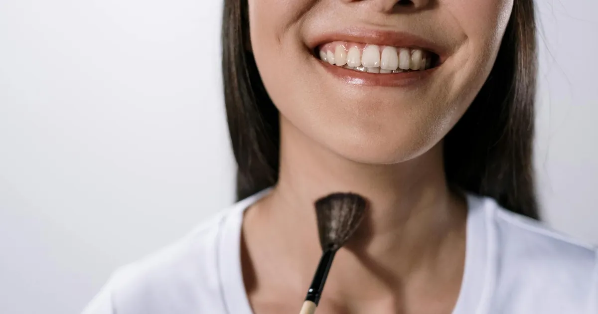 A smiling woman using a makeup brush in a studio on a white background.