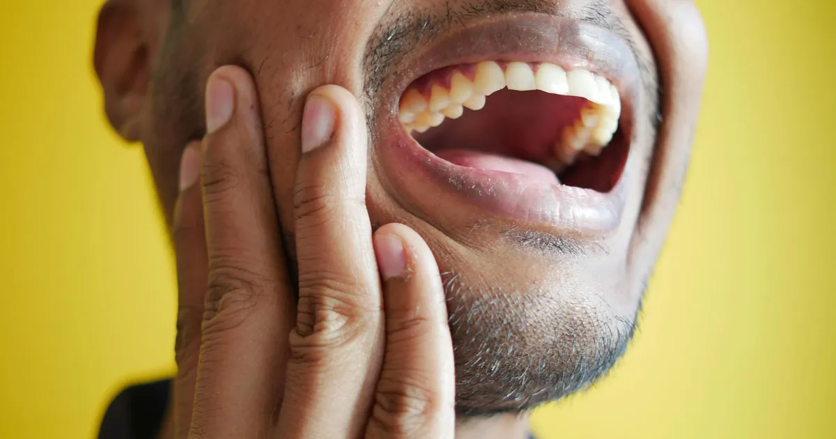 Close-up of a man holding his cheek, displaying a wide open mouth against a yellow background.