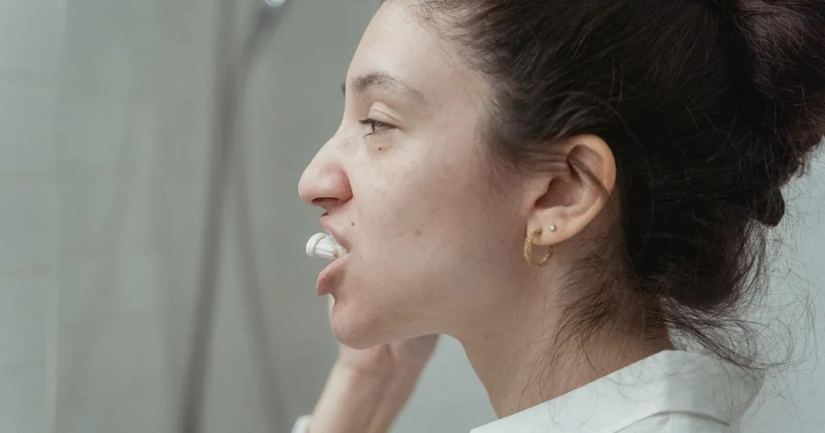 Side view of a woman with hair bun brushing teeth as part of her hygiene routine indoors.