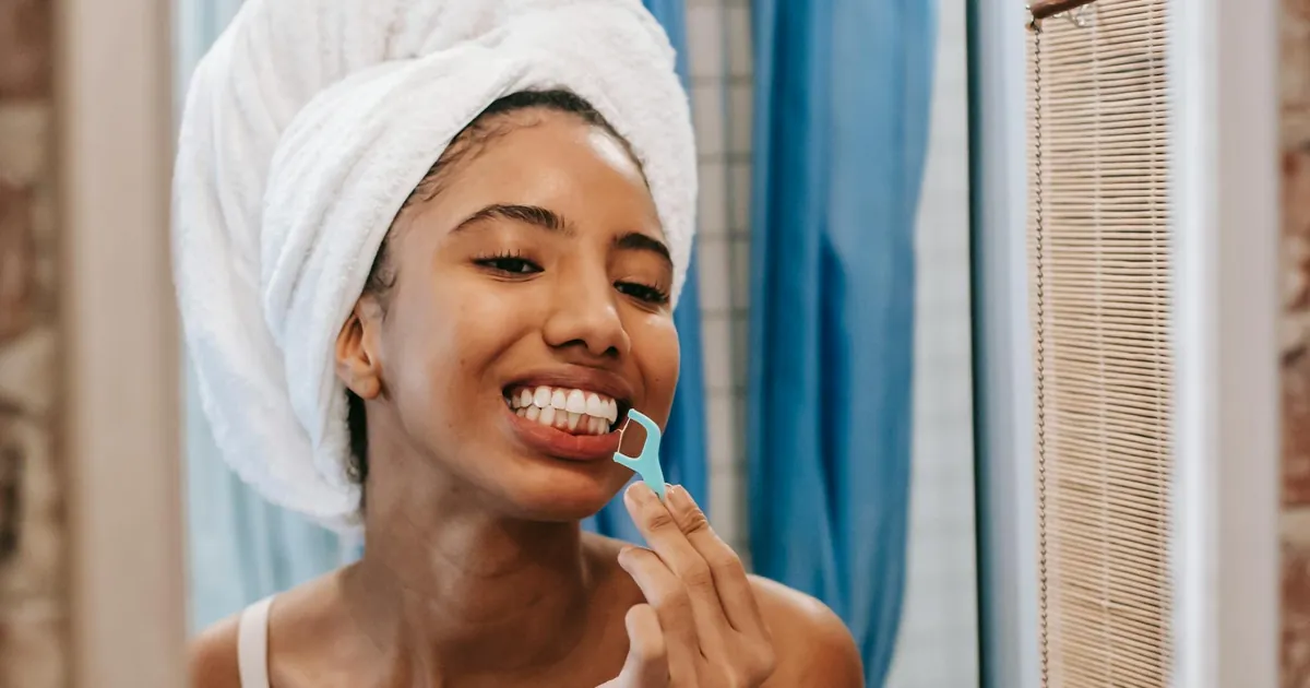 Young woman with towel on head flossing teeth in bathroom mirror, promoting hygiene and wellness.