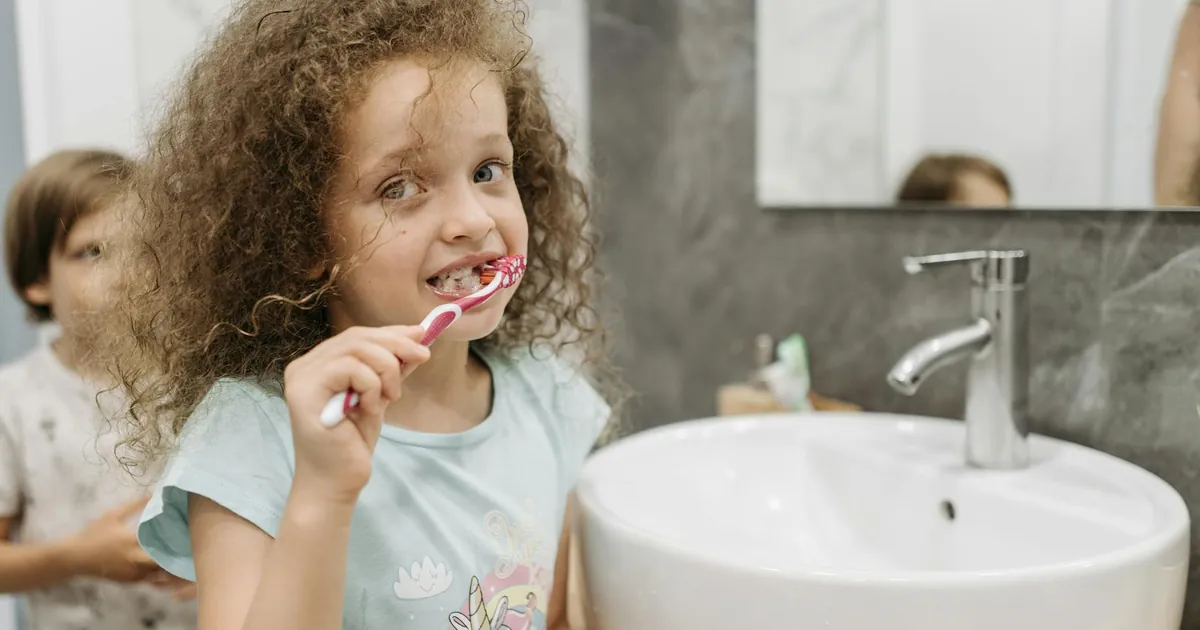 A young girl with curly hair brushes her teeth at a sink, promoting dental hygiene.