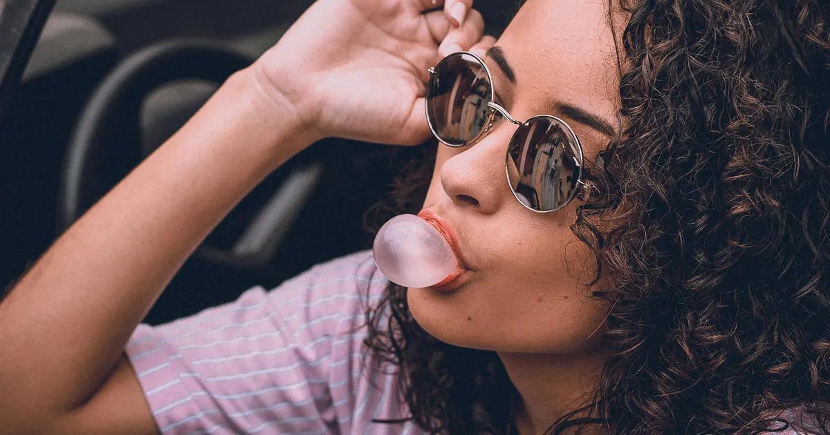 Woman with curly hair and sunglasses blowing gum in car, exuding style and confidence.